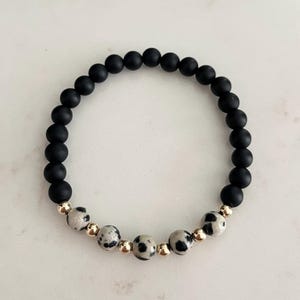 May include: A beaded bracelet featuring matte black and speckled gray beads, interspersed with gold-colored accent beads. The bracelet is circular and rests on a white surface. The bracelet is a piece of jewelry.