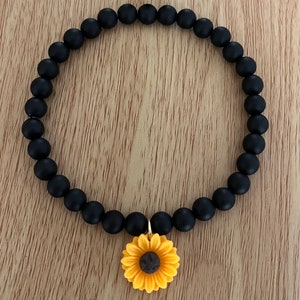 May include: Black beaded bracelet with a yellow and brown sunflower charm. The bracelet features matte black beads and a gold-colored clasp. The sunflower charm has yellow petals and a brown center.
