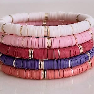 May include: Five stacked bracelets with gold accents. The bracelets are made of colorful ceramic beads in shades of white, pink, red, purple, and orange.
