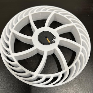 Pode incluir: Um objeto branco em forma de roda impresso em 3D com um disco preto central. A roda tem uma série de raios curvos e ranhurados e uma borda externa canelada. Um pequeno componente dourado é visível no disco preto.