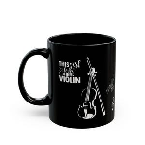 May include: Black ceramic mug featuring a white violin graphic, bow, musical notes, and the text "This girl loves her violin". The mug has a curved handle and a glossy finish.