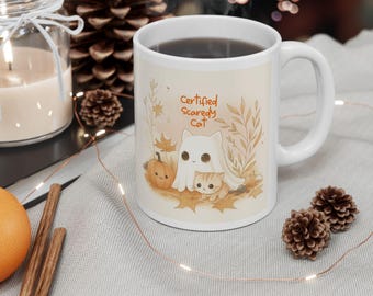 May include: White ceramic mug with a handle, featuring a "Certified Scaredy Cat" design. The design includes a ghost, a kitten, and a pumpkin, all surrounded by autumn leaves. The mug is filled with a dark liquid.