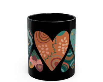 May include: Black ceramic mug featuring colorful heart designs. The hearts have various patterns, including floral, geometric, and abstract elements in shades of orange, green, and pink. The mug is a standard size for hot beverages.