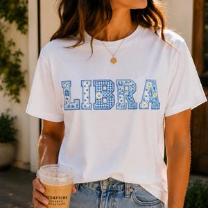 Puede incluir: Una camiseta blanca de manga corta con la palabra LIBRA impresa en el pecho en un patrón de cuadros vichy y flores en azul y blanco. La camiseta se combina con unos vaqueros de mezclilla azul claro con detalles desgastados en las rodillas.