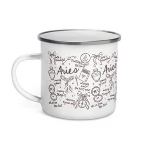 May include: White enamel mug with a silver rim and handle. The mug is decorated with Aries zodiac symbols and phrases like "bold is beautiful" and "trusting the spark". A charming gift for astrology enthusiasts.