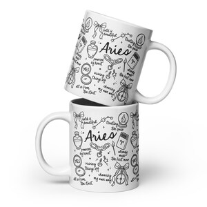 May include: Two white ceramic mugs stacked on top of each other. Each mug features a black and white Aries zodiac design with the word "Aries" and various illustrations and phrases like "bold is beautiful" and "choosing my own way."