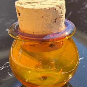 May include: A small, round, amber-colored glass jar with a wide, flat rim and a thick, cylindrical cork stopper. The glass has a smooth, translucent finish with a subtle iridescent sheen around the neck, and the cork has a textured surface.