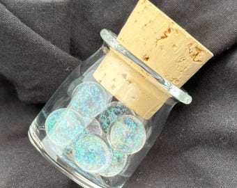 May include: A small, clear glass jar with a tapered neck and a thick, light brown cork stopper. Inside the jar are several round, flat glass cabochons that feature a shimmering, iridescent blue and green glitter finish that reflects light.