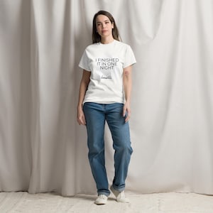 May include: A white t-shirt with the text "I FINISHED IT IN ONE NIGHT" above a small book illustration. The person is wearing blue jeans and white sneakers. The background is a white curtain.