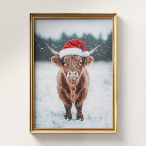 May include: A framed print featuring a brown Highland cow wearing a red Santa hat, standing in a snowy field. The print has a gold-colored frame and is set against a white wall. The cow has long horns and a fluffy coat.