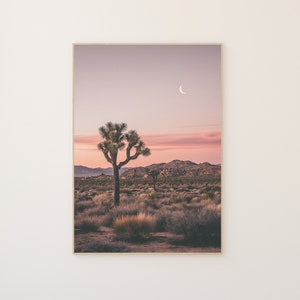 May include: A single Joshua tree stands in a desert landscape with a crescent moon in the sky. The sky is a soft pink and purple color, and the desert is covered in low-lying vegetation.