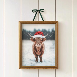 May include: A framed print featuring a Highland cow wearing a red Santa hat, set against a snowy backdrop. The print is in a gold frame and is suspended by a green ribbon, perfect for holiday decor.