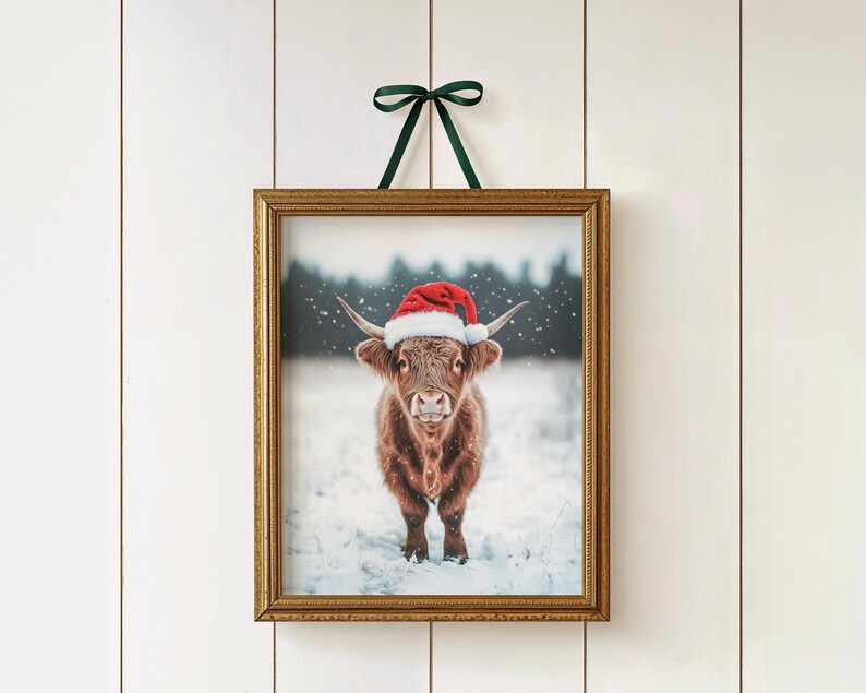 May include: A framed print featuring a Highland cow wearing a red Santa hat, set against a snowy backdrop. The print is in a gold frame and is suspended by a green ribbon, perfect for holiday decor.
