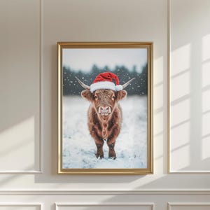 May include: A framed print featuring a Highland cow wearing a red and white Santa hat, standing in the snow. The cow has long, shaggy brown fur and curved horns. The background shows a snowy landscape with trees.