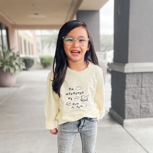 May include: A pale yellow long-sleeve sweatshirt featuring black text that reads ON WEEKENDS WE DIM SUM, accompanied by small line-art illustrations of dumplings and a steamer basket, paired with grey acid-wash denim jeans and clear-framed glasses.