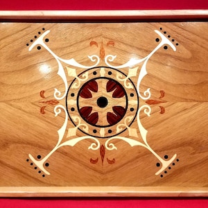 Puede incluir: Bandeja de madera para servir con un diseño decorativo incrustado. La bandeja presenta un fondo de veta de madera marrón claro y un patrón geométrico central en tonos marrón, blanco y negro. La bandeja es rectangular con bordes elevados.