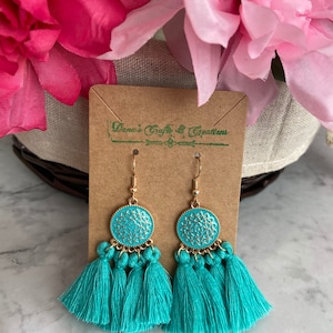 May include: Teal tassel earrings with gold-toned metal filigree and a turquoise blue center. The earrings are displayed on a brown card with the text "Dana's Crafts & Creations".