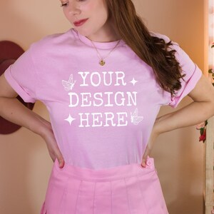 May include: A pink t-shirt with the text "YOUR DESIGN HERE" in white with butterfly graphics. The person is wearing a pink pleated skirt.