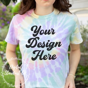 Può includere: Una t-shirt tie-dye con sfondo bianco e un motivo a spirale in tonalità di rosa, viola, blu e verde. Il testo "Your Design Here" è stampato in nero sul davanti della t-shirt.