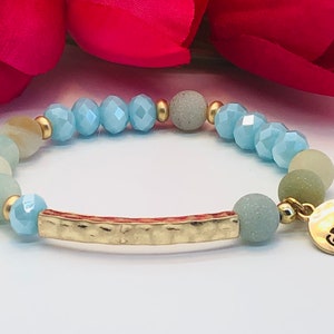 Peut inclure: Un bracelet en perles bleu clair avec un charme en forme de barre dorée et un charme en or gravé du mot "GRACE".