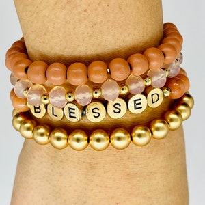 May include: A set of four bracelets featuring a gold beaded bracelet, a pink crystal bracelet, and two brown wooden bead bracelets. The pink crystal bracelet has gold beads and gold letter beads that spell out "BLESSED".