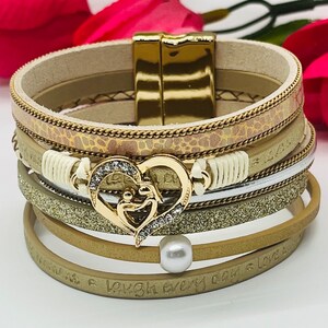 Peut inclure: Bracelet en cuir beige à plusieurs couches avec un charme en forme de cœur doré et un accent de perle. Le bracelet a un fermoir doré et les mots "laugh every day & love" sont gravés sur la couche inférieure.