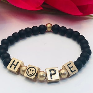 Peut inclure: Bracelet en perles noires avec des perles dorées et des breloques en lettres dorées qui épellent "HOPE" avec un breloque smiley.