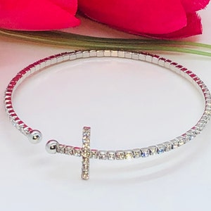 May include: A silver bangle bracelet with a clear crystal cross charm. The bracelet is made of small, square-shaped crystals.