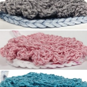 May include: Three crocheted circles in different colours: grey, pink, and teal. Each circle has a light blue crocheted border.