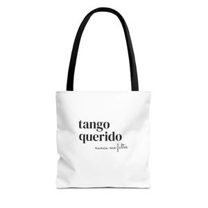 Puede incluir: Un bolso tote blanco con asas negras que presenta el texto impreso en negro "tango querido" en una fuente serif negrita, seguido del texto más pequeño en cursiva "nunca me faltes" colocado directamente debajo de la frase principal.