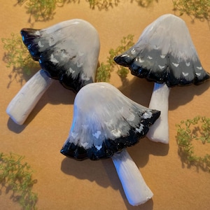 May include: Three ceramic mushroom ornaments with white stems and caps. The caps transition from white to grey and black, with white accents. The mushrooms are arranged on a tan surface with green moss.