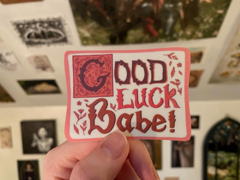 May include: A pink sticker with the text "Good Luck Babe!" in a red and black font with floral accents.
