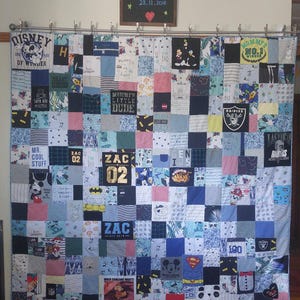 May include: A memory quilt constructed from various blue, white, and black fabric squares featuring graphic prints of Disney characters, sports logos, and text such as Zac 02, Mummy's No. 1 Winner, and Mr. Cool Stuff, hanging from a metal rod.