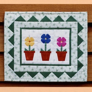 Può includere: Un modello di blocco di trapunta in carta a pezzi per una piccola trapunta con tre vasi di fiori con fiori gialli, blu e rosa. Il blocco di trapunta misura 11 pollici per 13 pollici ed è intitolato "Posy Pots".