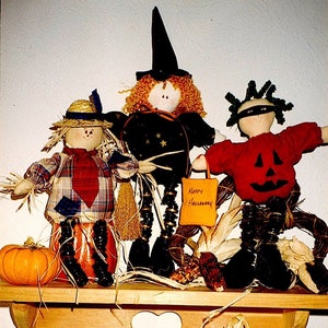 Pode incluir: Três bonecas de tecido com tema de Halloween: uma bruxa com um chapéu preto e um vestido com estampa de estrelas, um espantalho com uma camisa xadrez e um chapéu de palha, e uma abóbora com uma camisa vermelha e uma máscara preta para os olhos. As bonecas estão dispostas em uma prateleira de madeira com decorações de outono.