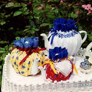 Peut inclure: Trois chauffe-théières en tissu dans un panier en osier blanc. Les chauffe-théières sont en tissu avec différents motifs et couleurs. L'un est jaune avec des oiseaux bleus, l'autre est rouge avec des fleurs blanches et le dernier est blanc avec un tissu à carreaux bleus et blancs.