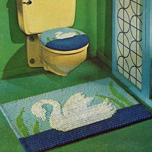 Op de afbeelding: Een vintage brei- en haakpatroonboek met een badkamerscène met een toilet, een toiletbrilhoes en een badmat, allemaal gehaakt in een zwanenontwerp. De omslag van het boek bevat de tekst "Knit and Crochet with Aunt Lydia's Heavy Rug Yarn" en de prijs "29c".