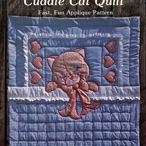 Könnte beinhalten: Eine blaue und weiße Gingham-Steppdecke mit einer rosa und weißen Applikation eines Katers mit Schleife. Die Steppdecke ist mit einem Rautenmuster gesteppt. Der Text "Cuddle Cat Quilt" und "Fast, Fun Applique Pattern" befindet sich oben im Bild. Der Text "The Gingham Goose" befindet sich unten im Bild. Der Text "Patterns & Pretties" befindet sich unter "The Gingham Goose". Der Text "Designed and Created by Gwen Andrews © 1979, Gwen Andrews" befindet sich unter "Patterns & Pretties". Der Text "V.I.P. DIV. OF CRANSTON PRINT WORKS CO. 1412 Broadway * New York, N.Y. 10018" befindet sich unten im Bild.