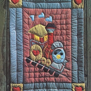 Op de afbeelding: Een quiltpatroon voor een 'Chuggy Choo-Choo Quilt' met een rode, gele en blauwe trein-applique op een rood-witte ruitjesachtergrond. Het patroon is ontworpen door Gwen Andrews en is afkomstig uit de collectie 'The Gingham Goose'.