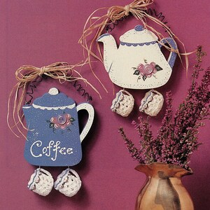 May include: Two wooden tea pot and coffee pot plaques with crocheted accents hanging on a purple wall. The coffee pot is blue with white trim and says "Coffee". The tea pot is white with blue trim and floral accents.