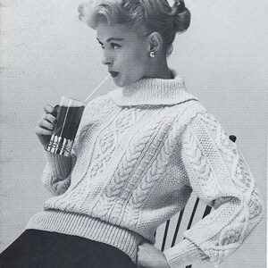 May include: A woman wearing a white cable knit sweater with a turtleneck. She is holding a glass with a straw and a dark liquid inside. She is sitting in a chair with a black and white pattern.