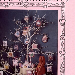Pode incluir: Um conjunto de 12 padrões de ponto cruz para ornamentos de Páscoa. Os padrões apresentam uma variedade de desenhos, incluindo coelhos, pintinhos, ovos e flores. Os padrões são impressos em um fundo rosa com uma borda quadriculada preta e branca. O texto "PENSTITCH EASTER ORNAMENTS" é impresso na parte superior da página. O texto "MUMM'S THE WORD" é impresso na parte inferior da página.