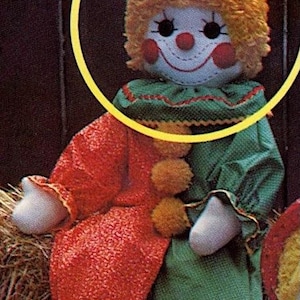 May include: A handmade clown doll with a red and green outfit. The clown has a red and green hat, orange hair, and a painted face with a smile. The doll is sitting on a hay bale.