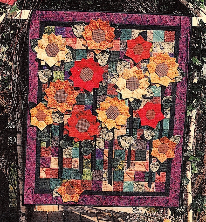 K&ouml;nnte beinhalten: Ein farbenfroher Quilt mit Patchwork-Muster aus verschiedenen Stoffen in Rot-, Orange-, Gelb-, Gr&uuml;n-, Blau- und Violettt&ouml;nen. Der Quilt ist mit mehreren aufgen&auml;hten Sonnenblumen in verschiedenen Braunt&ouml;nen, Orange- und Rott&ouml;nen verziert.