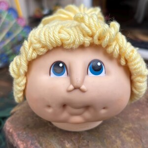 May include: A Cabbage Patch Kid doll head with yellow yarn hair and blue eyes. The doll has a rosy complexion and a sweet smile.