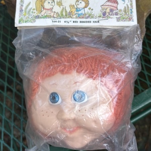 Puede incluir: Una cabeza de muñeca de plástico con cabello rojo trenzado y ojos azules. La cabeza de la muñeca está empaquetada en una bolsa de plástico transparente con una tarjeta blanca que dice "Baby Toothie" y "4 1/2" Red Braided Hair".