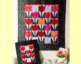 May include: A pattern for miniature quilts featuring tulip designs. The main quilt measures 14 inches by 16 inches, with a smaller version at 6.75 inches by 8 inches. The quilts showcase red, pink, and orange tulips on a white background, framed by a dark green border.