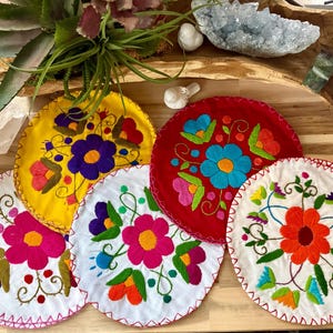 May include: A collection of colorful, embroidered round placemats. Each mat features a different floral design in vibrant colors like yellow, red, white, and pink. The edges are finished with a contrasting stitch.