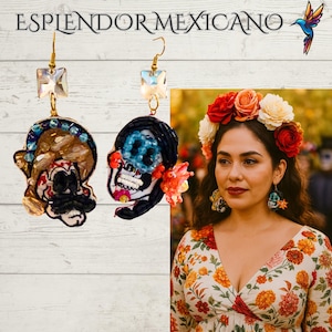 May include: Embroidered skull earrings with crystal accents. One earring features a skull with a black mustache and a beaded headdress. The other earring has a blue skull with black hair and a floral accent. The earrings are gold-toned and have a square crystal at the top. The text "ESPLENDOR MEXICANO" is at the top.