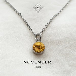 May include: A silver chain necklace with a small pendant featuring a round topaz gemstone. The pendant is a light yellow colour. The text "NOVEMBER Topaz" is written below the pendant.